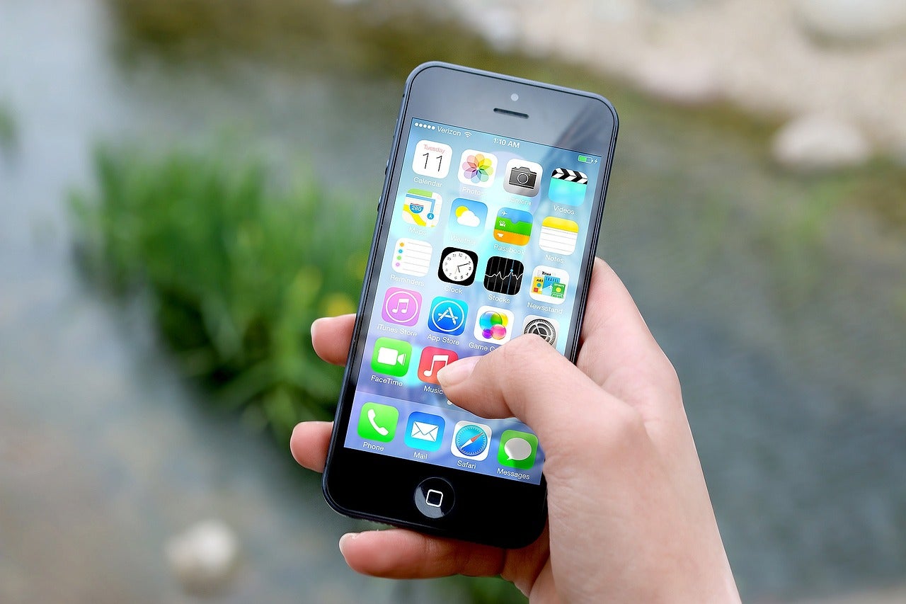 human right hand holding iPhone with apps on screen and thumb pressing icon; background of a stream with rocks and stones.