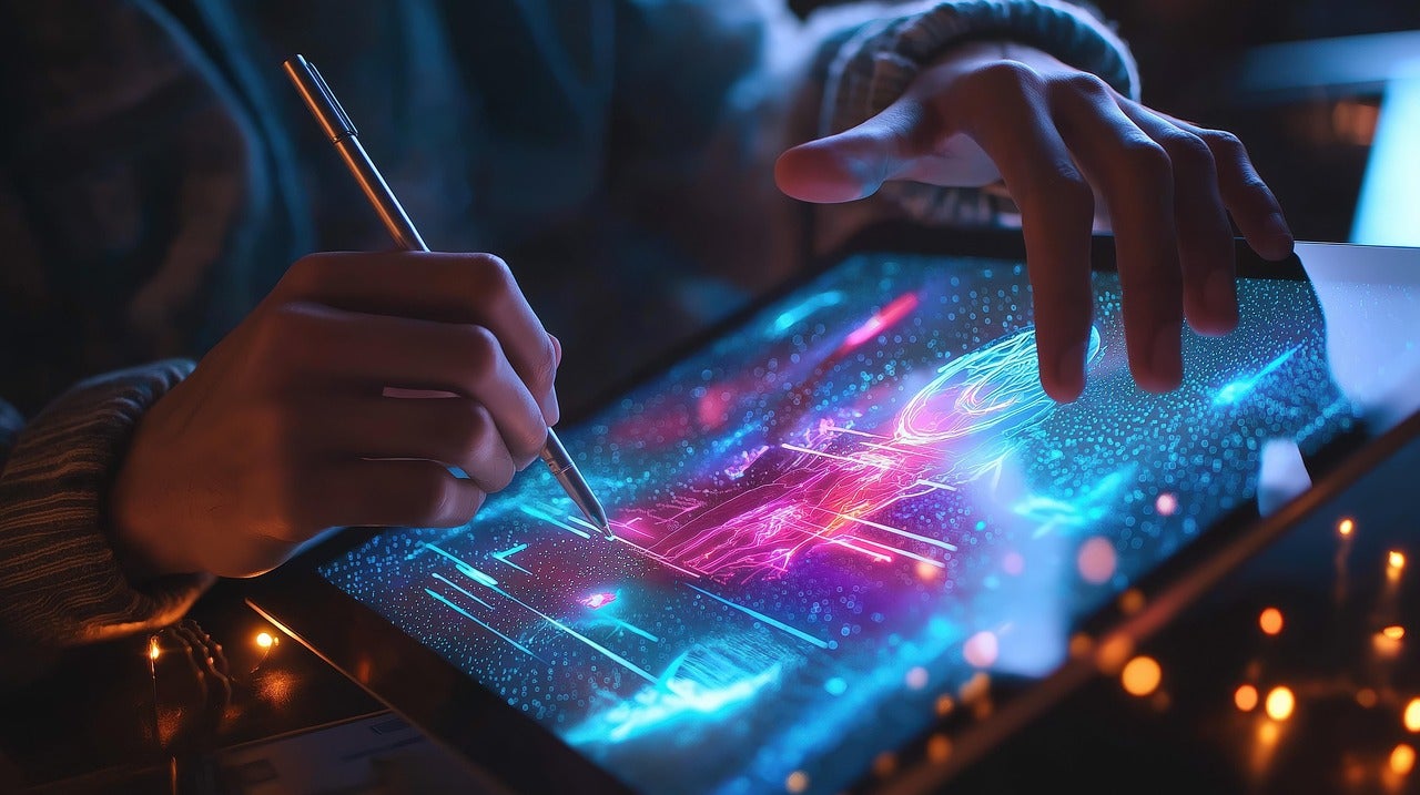 User working in large touch screen tablet in dim lighting. The screen glows hot pink and luminescent turqoise with the users hands touching the screen and holding a stylus.