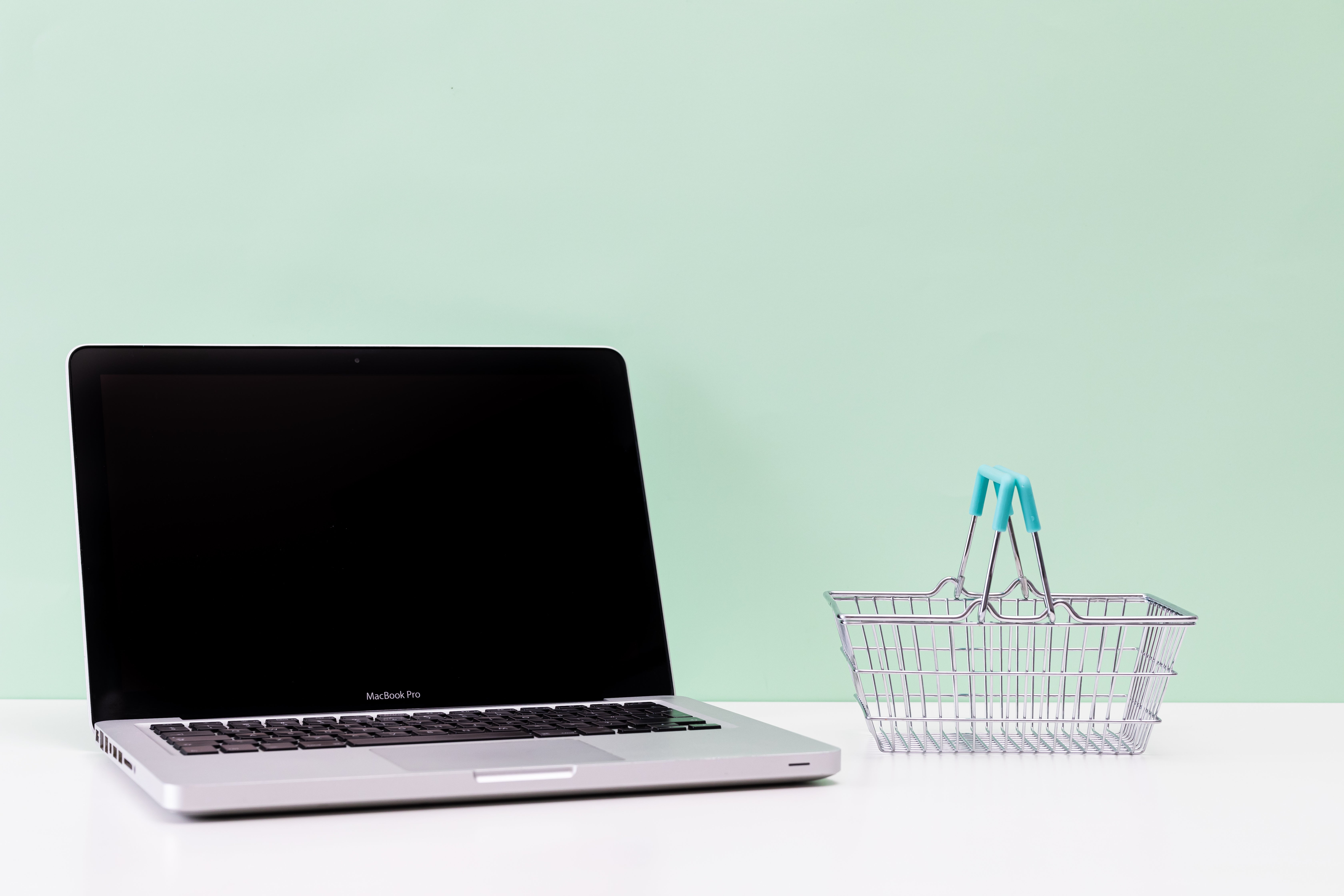 Laptop with small basket image open laptop on a white surface next to a miniature metal basket with blue handles with a sage green background from Kaboompics.com