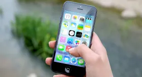 human right hand holding iPhone with apps on screen and thumb pressing icon; background of a stream with rocks and stones.