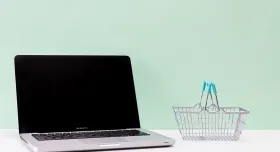 open laptop on a white surface next to a miniature metal basket with blue handles with a sage green background from Kaboompics.com