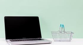 open laptop on a white surface next to a miniature metal basket with blue handles with a sage green background from Kaboompics.com