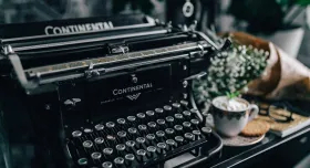 early era black typewriter on table seen at an angle. A cup of coffee and a bouquet of babies breath sit adjacent to the typewriter. From Kaboompics.com